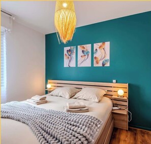 A modern bedroom with teal walls, a wooden bed frame, a grey knitted throw, and three wall-mounted paintings.