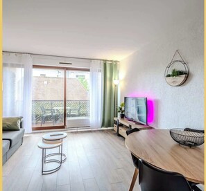 A modern living room with a wooden dining table, a TV on a stand, and a balcony with a view of the neighborhood.