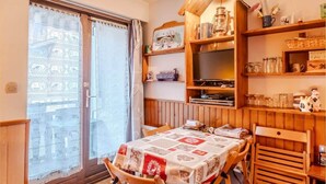 Un rincón comedor acogedor con una mesa preparada para dos, sillas de madera, una estantería con diversos objetos y un ventanal con cortinas estampadas.