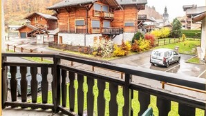Una cabaña de madera con balcón, un coche aparcado delante y montañas al fondo.