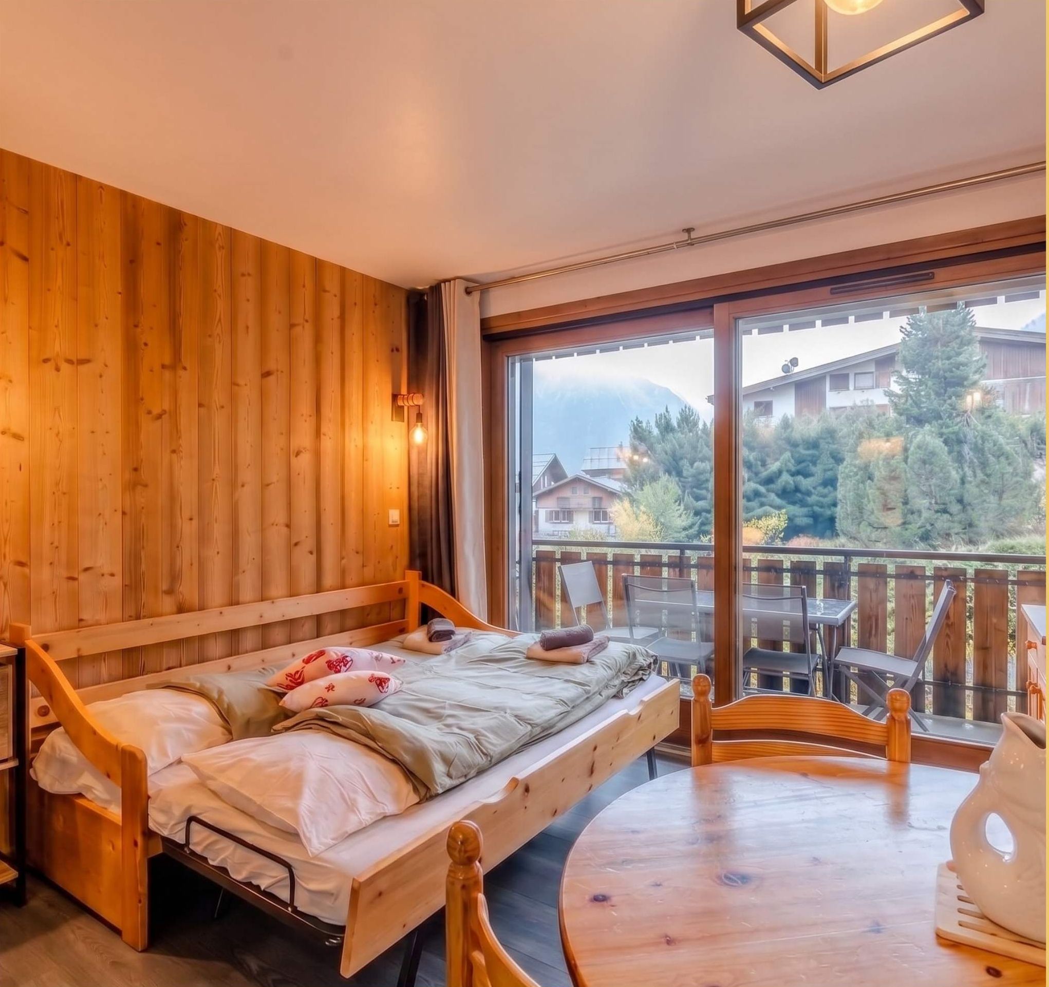 A wooden room with a bed, a table, and a view of houses and trees.