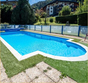 A rectangular swimming pool with a blue interior, surrounded by a grassy area and a pebble-tiled deck. - Le Paradisio I Studio I Morzine (Morzine)