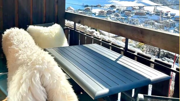 Een balkon met een houten hekwerk, een witte, pluizige stoel en een metalen tafel.