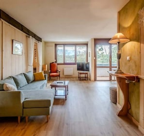 Un salon spacieux comprenant un canapé, un fauteuil et un téléviseur, ainsi que des planchers en bois et de grandes fenêtres donnant sur l’extérieur.