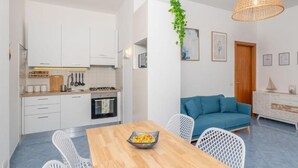 A modern kitchen with white cabinets, a dining table with a bowl of fruit, and a blue sofa in the living area. - Villa Marea, Praiano (Praiano)