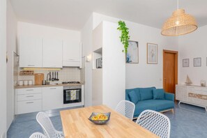 Eine moderne Küche mit weißen Schränken, einem Esstisch mit Obstschale und einem blauen Sofa im Wohnbereich.