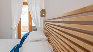 A bedroom with a wooden headboard, white bedding, and a view through a window with white curtains. - Villa Marea, Praiano (Praiano)
