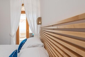 Ein Schlafzimmer mit einem hölzernen Kopfteil, weißer Bettwäsche und Blick durch ein Fenster mit weißen Vorhängen.