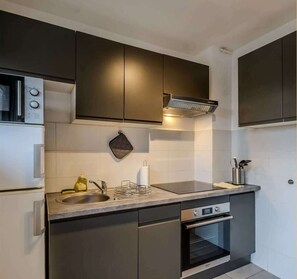 A modern kitchen with dark cabinetry, a stainless steel range hood, a built-in oven, and a sink with a dish rack. - Little cocoon I Studio I Annemasse (Annemasse)