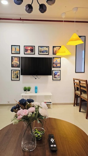 Un salon avec un téléviseur à écran plat fixé au mur, entouré d’affiches de voyage encadrées. Une table ronde en bois, sur laquelle se trouvent un vase de fleurs et une télécommande, est placée au premier plan.