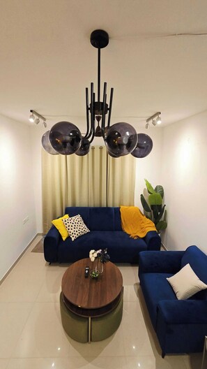 A modern living room with a blue sofa set, a round coffee table with a vase of flowers, and a black chandelier.