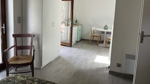 Quarto com guarda-roupa, uma cadeira, uma mesinha com um vaso e uma cozinha compacta ao fundo.