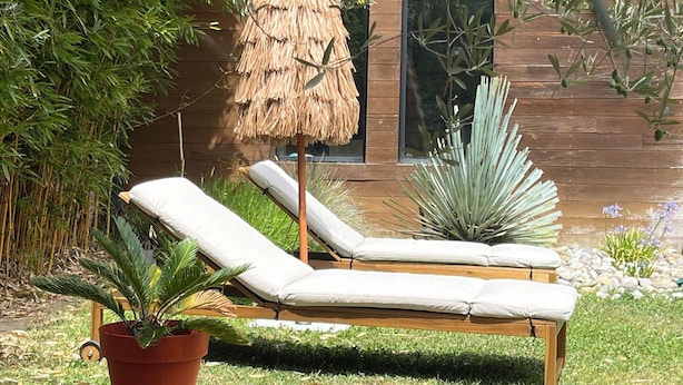 Um deck de madeira com duas cadeiras de lounge e um guarda-sol de palha, rodeado por vegetação exuberante e um edifício de madeira ao fundo.