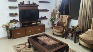 A living room with a TV, wooden entertainment unit, two armchairs, a coffee table, and decorative plants. - Shahed Apartment 3-bedroom apartment in charming Amman with comfort (Amman)