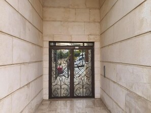 Corredor com porta dupla adornada com detalhes em metal, que dá para área externa com carros estacionados e rua à vista.
