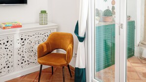 A room with a white wall, a flat-screen TV, a white cabinet with decorative patterns, a mustard-colored chair, a teal-striped curtain, and a glass door leading to an outdoor area with a table and potted plants.
