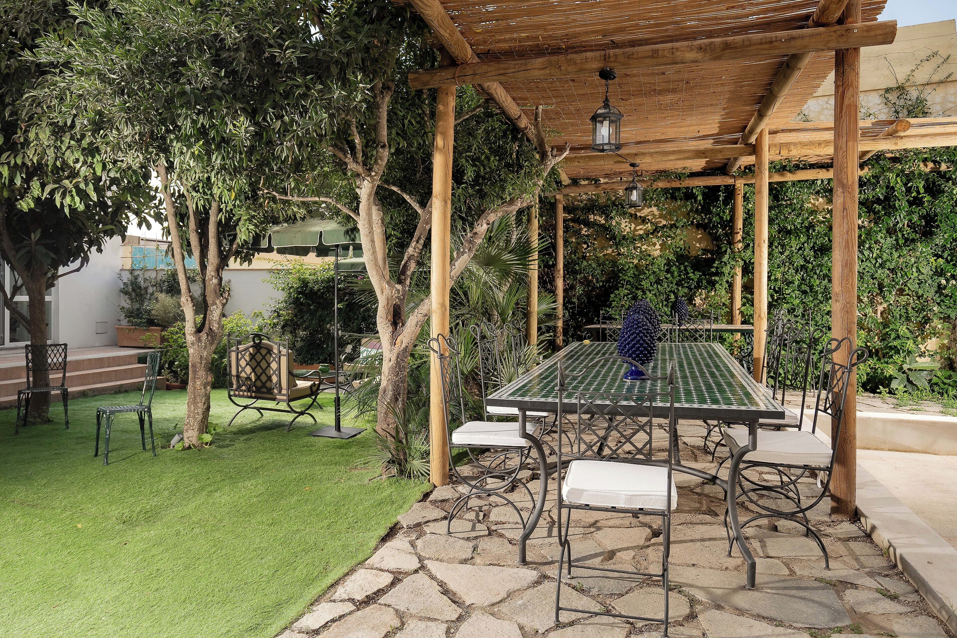 A covered outdoor seating area with a table and chairs, surrounded by trees and a well-maintained lawn.