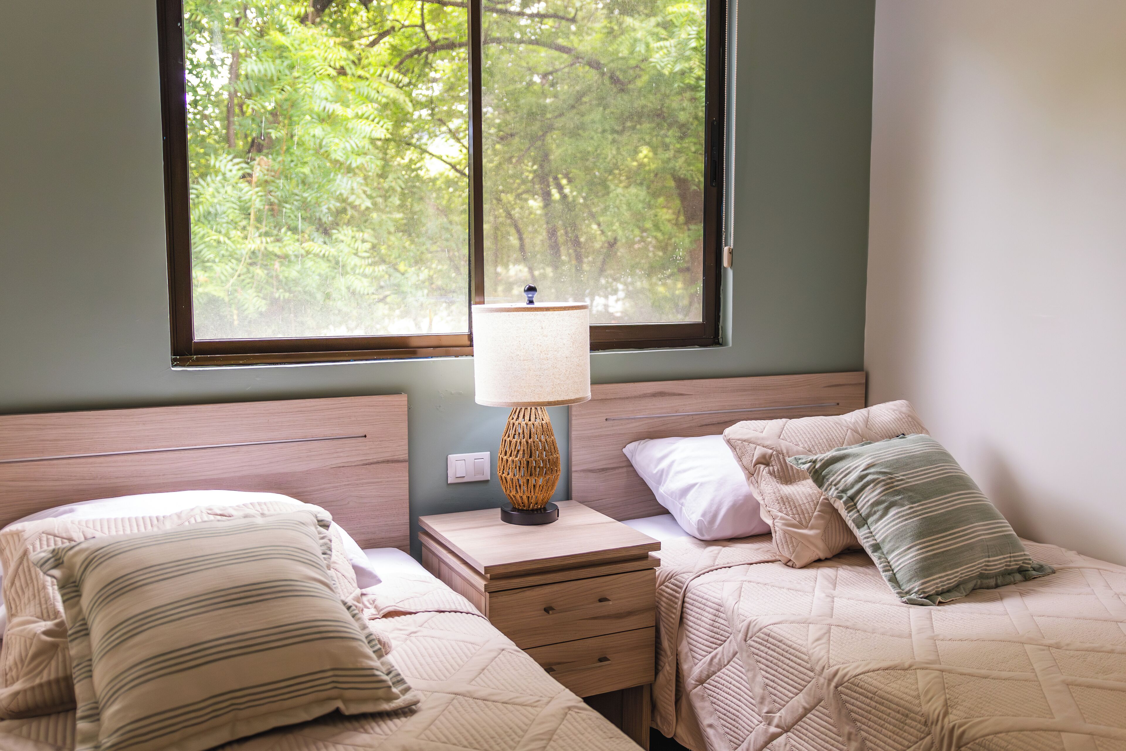 A bedroom with two beds, a nightstand with a lamp, and a window with a view of trees. - Tropical 2nd floor Condo with Rooftop (Playas Del Coco)