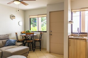 A compact living space with a sofa, dining table, chairs, a ceiling fan, a window with a view of greenery, and a kitchen area with a sink and cabinets. - Tropical 2nd floor Condo with Rooftop (Playas Del Coco)