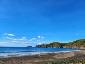 A beach with clear blue water, a sandy shore, and green hills in the background. - Tropical 2nd floor Condo with Rooftop (Playas Del Coco)