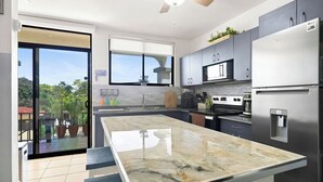 A modern kitchen with a large island, stainless steel appliances, and a sliding glass door leading to a balcony with potted plants. - Casa Elevación | Ocean-View Rooftop Retreat | Pool | Fiber WiFi (Playas Del Coco)