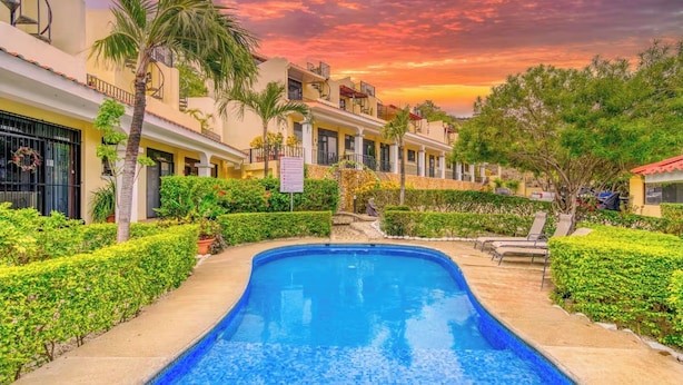 A swimming pool surrounded by well-maintained hedges and palm trees, with a backdrop of a building complex and a sunset sky. - Casa Elevación | Ocean-View Rooftop Retreat | Pool | Fiber WiFi (Playas Del Coco)