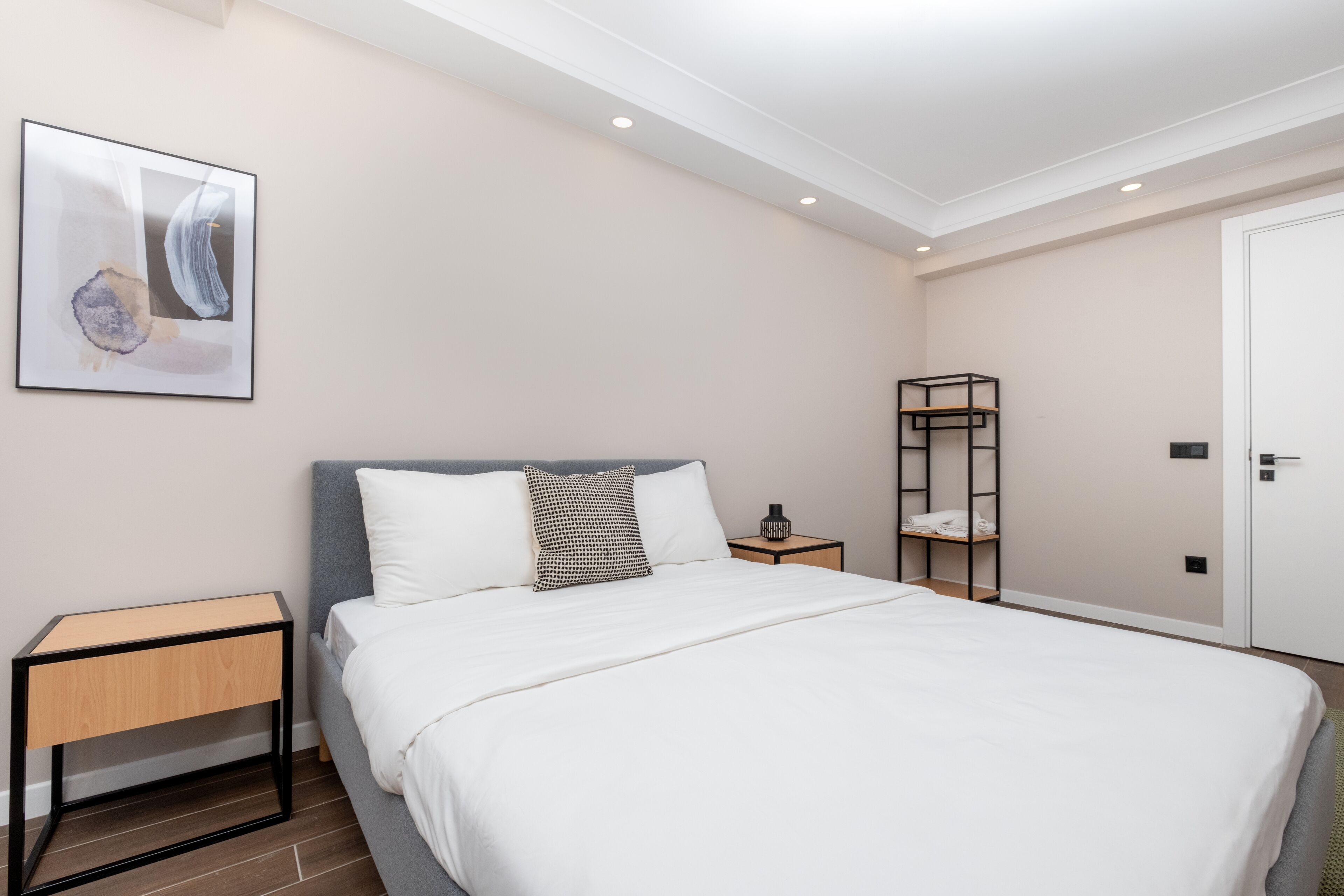 A neatly arranged bedroom with a bed, a bedside table, a nightstand, a wall-mounted shelf, and a door.