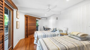 Quarto com duas camas, ventilador de teto, escrivaninha com cadeira e janela com persianas.