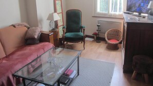 A living room with a sofa, a glass coffee table, a green armchair, a wicker chair, a lamp, a painting, and a television. - Village house with 2 bedrooms, garden and outdoor swimming pool (Distré)