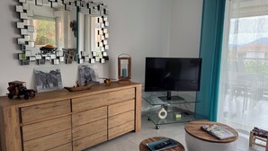 Una sala de estar moderna con un aparador de madera, una mesa de centro de vidrio, un taburete redondo de madera, un televisor y un espejo fijado a la pared.