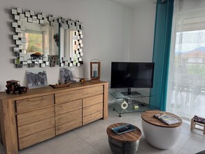Un salon moderne avec un meuble en bois, une table basse en verre, un tabouret rond en bois, un téléviseur et un miroir fixé au mur.
