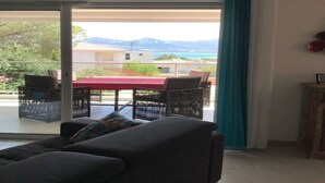 Habitación con sofá, mesa y sillas, puerta corrediza de vidrio que da a un espacio exterior con mesa y sillas, y vistas a montañas y agua.