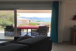 Une pièce comprenant un canapé, une table et des chaises, une porte-fenêtre coulissante donnant sur un espace extérieur avec une table et des chaises, et offrant une vue sur les montagnes et l’eau.