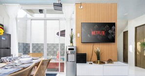 A modern living room with a flat-screen TV displaying 'NETFLIX,' a dining area with a table set for four, and a water dispenser.