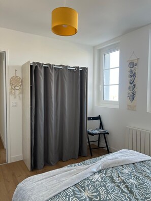 A bedroom with a bed, a chair, a window, a curtain, a wall hanging, and a hanging light fixture.