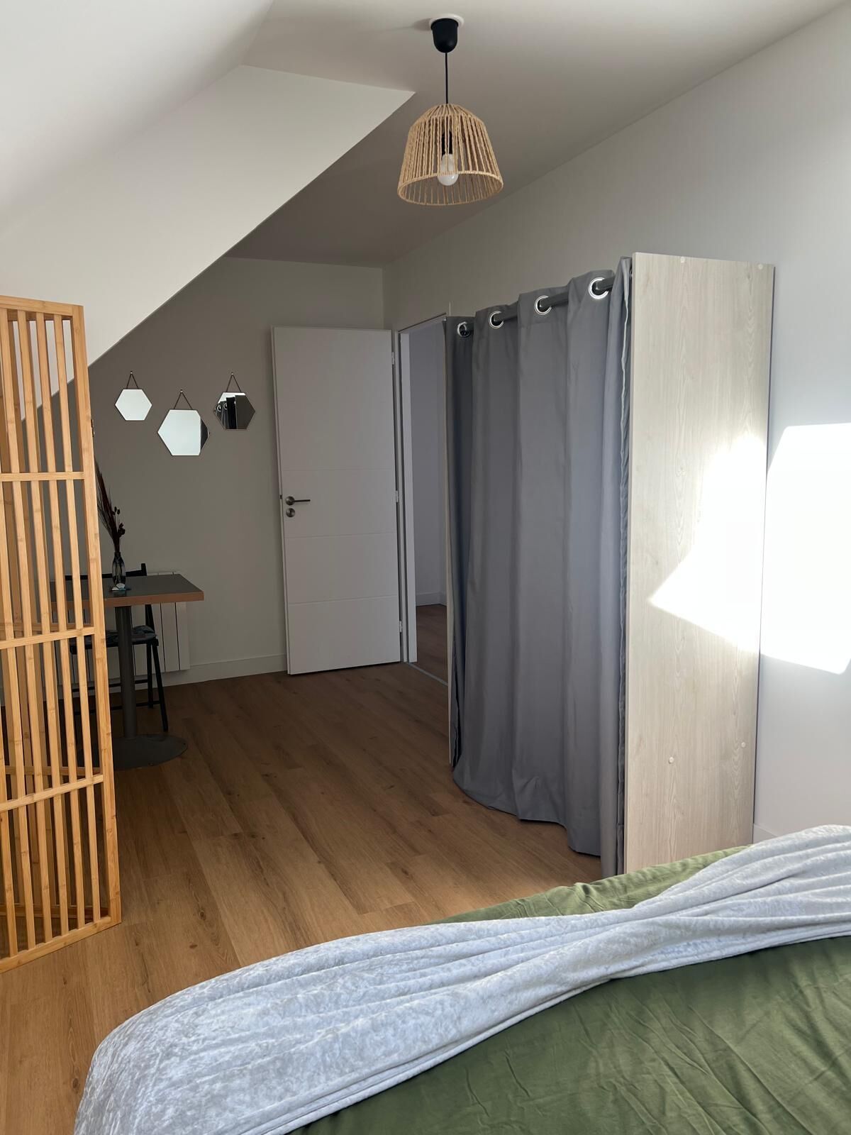 A room with a bed, a wooden partition, a desk with a chair, a hanging light fixture, and a door leading to another room.