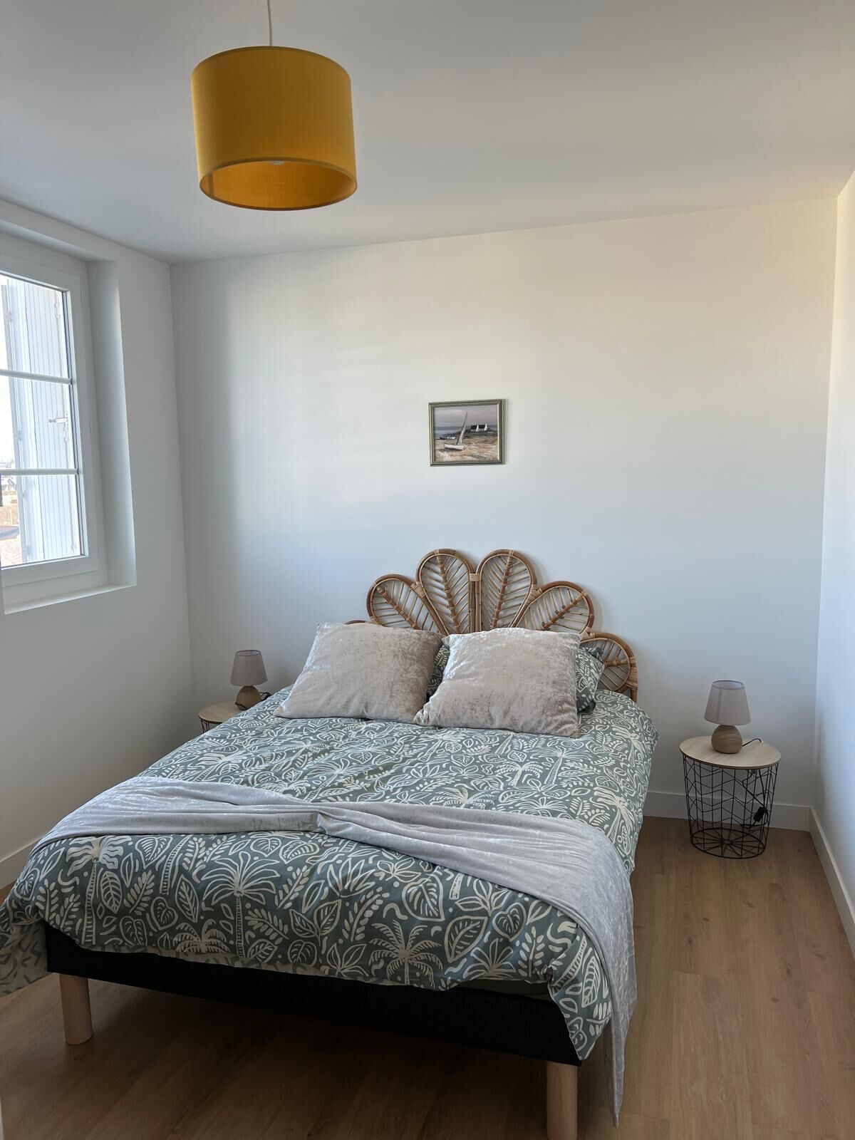 Un dormitorio con cama, lámpara colgante, una mesita redonda, un cuadro en la pared y una ventana.