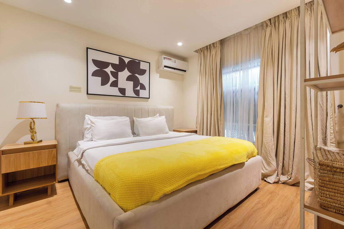A bedroom with a bed, a nightstand, a lamp, a wall-mounted air conditioner, and a window with curtains.