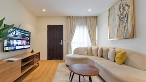 A modern living room with a flat-screen TV, a wooden entertainment unit, a beige sofa with cushions, a round coffee table, and a large abstract painting on the wall. - Aima Apartments  (Lekki)