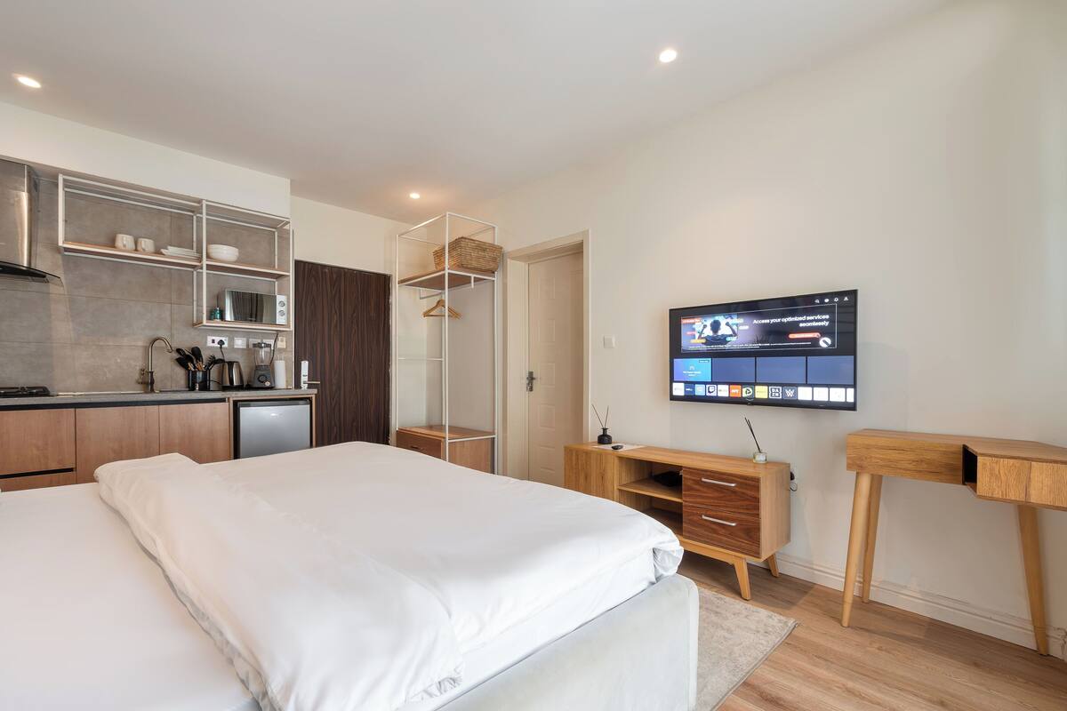 A modern bedroom with a bed, a desk, a TV, and a kitchenette.