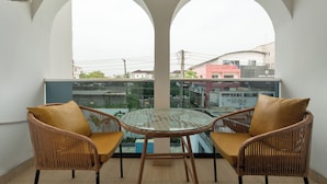 A balcony with two wicker chairs and a glass table, overlooking a cityscape with buildings and a pink hotel. - Aima Apartments  (Lekki)