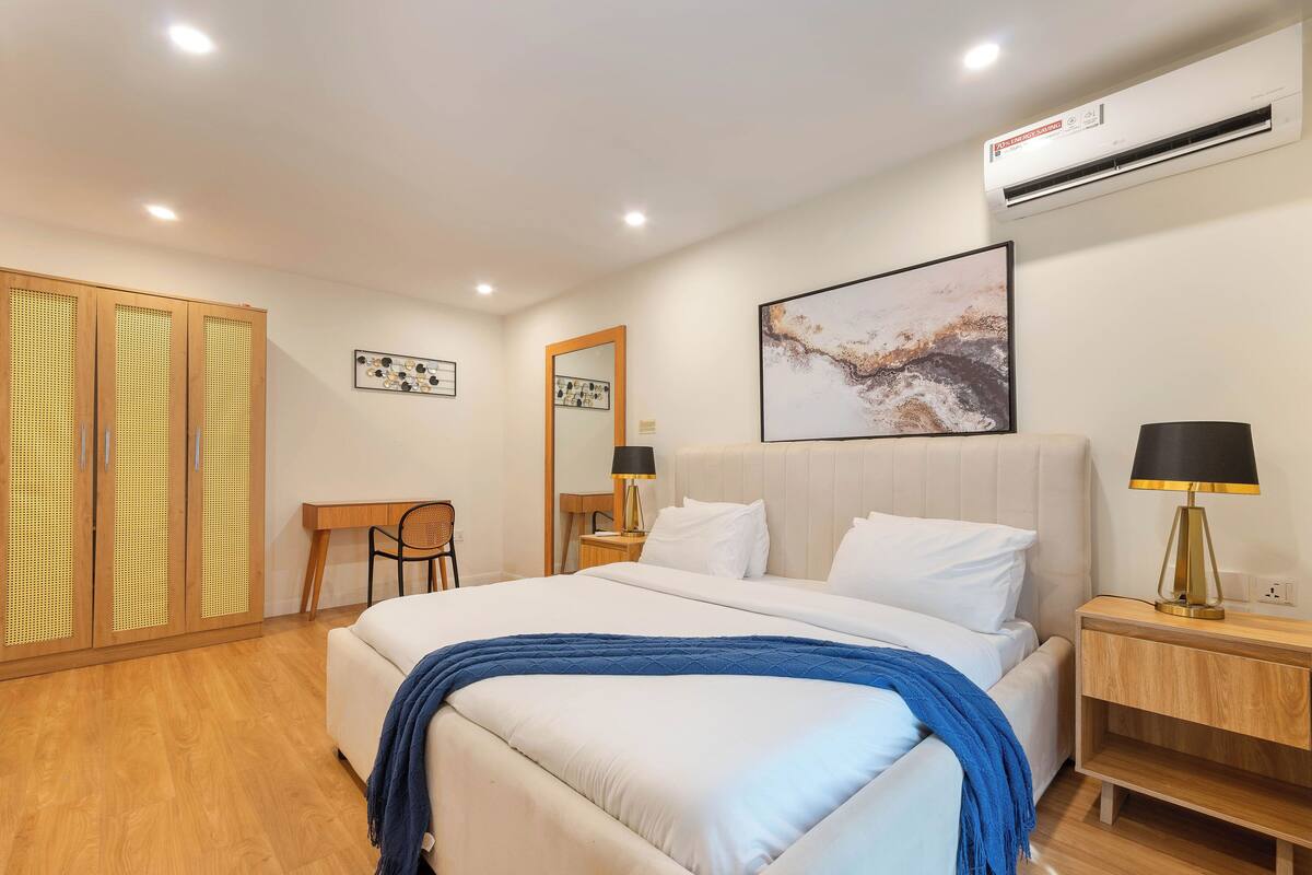 A modern hotel room with a large bed, wooden wardrobe, desk, and a mirror.