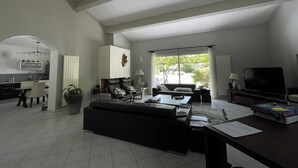 A spacious living room with a black sofa, a dining table, and a TV. There is a kitchen area with a fridge and cabinets. - Near Montpellier - Country house 8 to 10 persons (Saint-Clément-de-Rivière)