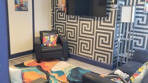 Una stanza con un muro decorato, un televisore a schermo piatto sopra una poltrona in pelle nera, un tappeto colorato e un divano con un cuscino decorato.