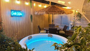 Un espace piscine extérieur agrémenté d’un panneau lumineux au néon, de meubles en osier et d’un parasol en chaume.