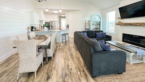 A spacious living area with a dark gray sofa, wicker chairs, a dining table, and a kitchen in the background. - Unit 028 - Sea Goodness (Dauphin Island)