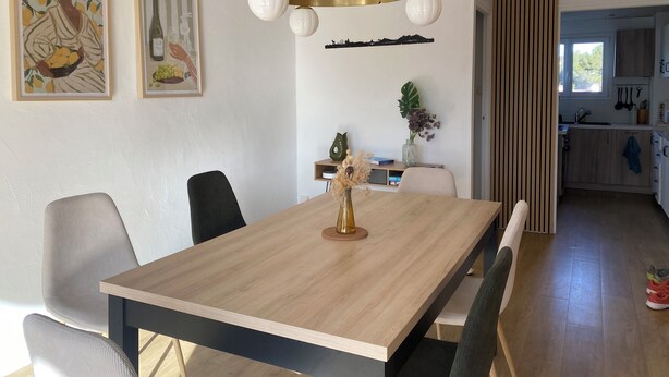 Un espace repas moderne avec une table en bois, des chaises noires et un luminaire suspendu. Des œuvres d’art encadrées ornent le mur, et on aperçoit une cuisine au fond.
