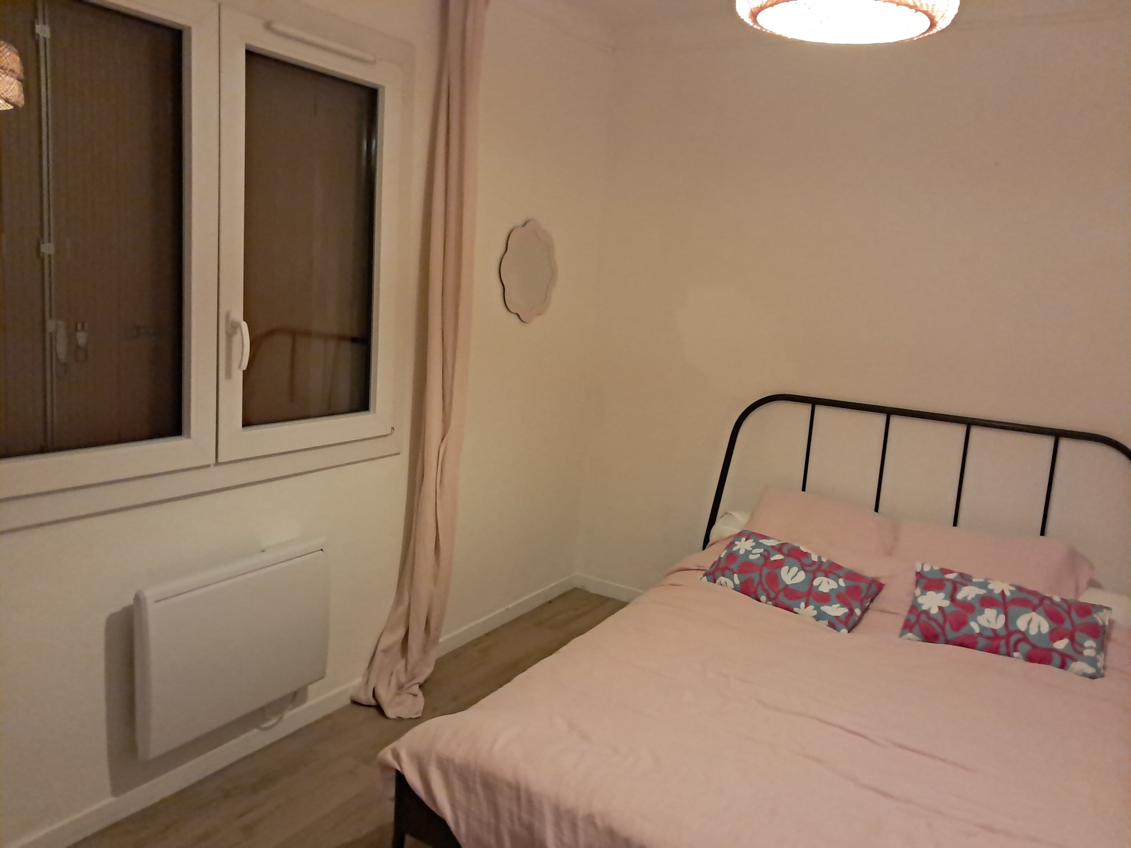 A bedroom with a bed, a window with curtains, a radiator, and a wall-mounted light fixture.