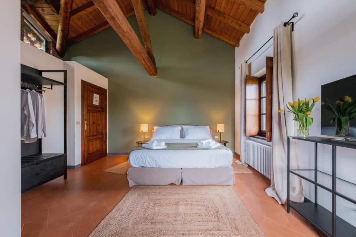 A bedroom with a large bed, wooden beams, a radiator, and a TV on a black stand.