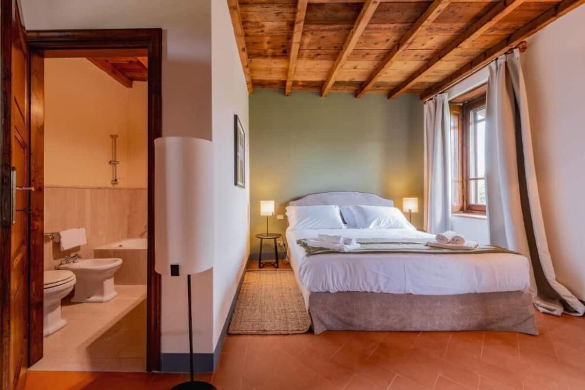 A bedroom with a wooden ceiling, a bed with white linens, a bedside table with a lamp, and a window with curtains.